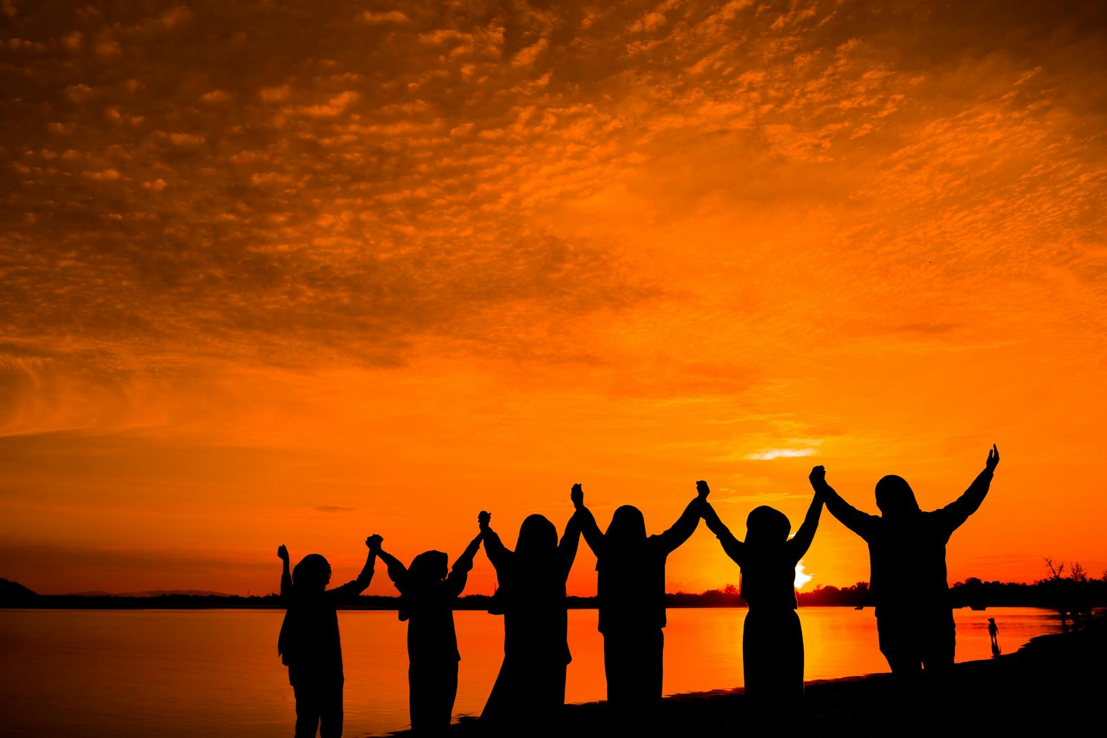 A line of people in silhouette holding hands and raising their arms toward a glowing orange sunset over calm water.